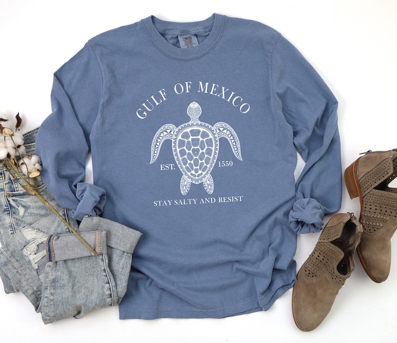 Op de afbeelding: Een blauw longsleeve t-shirt met een witte afbeelding van een zeeschildpad en de tekst "Gulf of Mexico Est. 1550 Stay Salty and Resist".