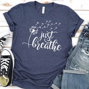 Op de afbeelding: Een marineblauw t-shirt met een witte paardenbloem afbeelding en de tekst "just breathe".