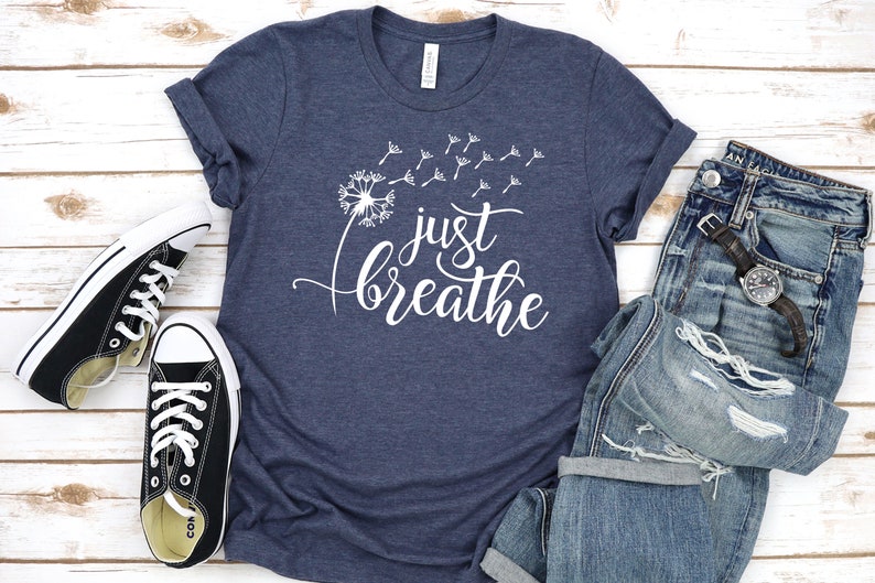 Op de afbeelding: Een marineblauw t-shirt met een witte paardenbloem afbeelding en de tekst "just breathe".
