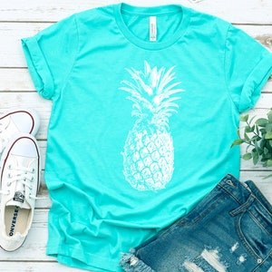 Pode incluir: Uma camiseta turquesa com um gr&aacute;fico de abacaxi branco. A camiseta est&aacute; colocada sobre uma superf&iacute;cie de madeira branca com um par de t&ecirc;nis brancos, um par de cal&ccedil;as jeans azuis e uma pequena planta verde.