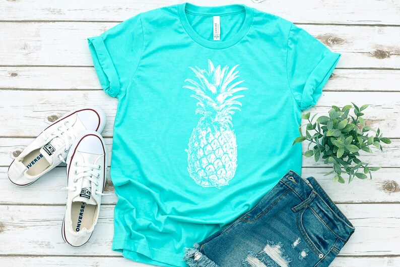 Pode incluir: Uma camiseta turquesa com um gr&aacute;fico de abacaxi branco. A camiseta est&aacute; colocada sobre uma superf&iacute;cie de madeira branca com um par de t&ecirc;nis brancos, um par de cal&ccedil;as jeans azuis e uma pequena planta verde.
