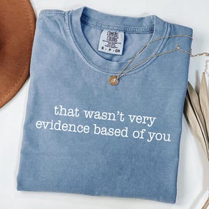 May include: A blue Comfort Colors t-shirt with the phrase "that wasn't very evidence based of you" in white. A gold necklace with a pendant is draped across the shirt. A brown hat and dried plant are in the background.