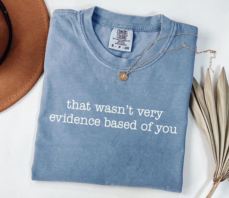 May include: A blue Comfort Colors t-shirt with the phrase "that wasn't very evidence based of you" in white. A gold necklace with a pendant is draped across the shirt. A brown hat and dried plant are in the background.