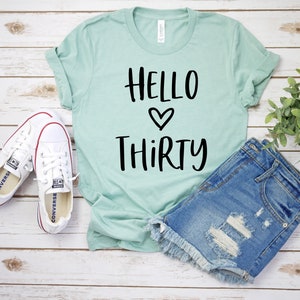 May include: A light green t-shirt with the text "Hello Thirty" in black lettering with a heart symbol between the words. The t-shirt is laying on a white wooden surface with a pair of white sneakers and a pair of blue denim shorts.