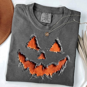 May include: A charcoal gray t-shirt featuring a distressed jack-o'-lantern face graphic with orange fabric showing through the frayed cutouts. The inner neck label reads Comfort Colors, 100% cotton, made in Honduras, size S-P-CH.