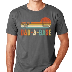 Op de afbeelding: Donkergrijs t-shirt met een retro design. Het ontwerp toont een floppy disk graphic met de tekst "I KEEP ALL MY DAD JOKES IN A DAD-A-BASE" in oranje, geel, blauwgroen en wit. Het t-shirt heeft een ronde hals en korte mouwen.