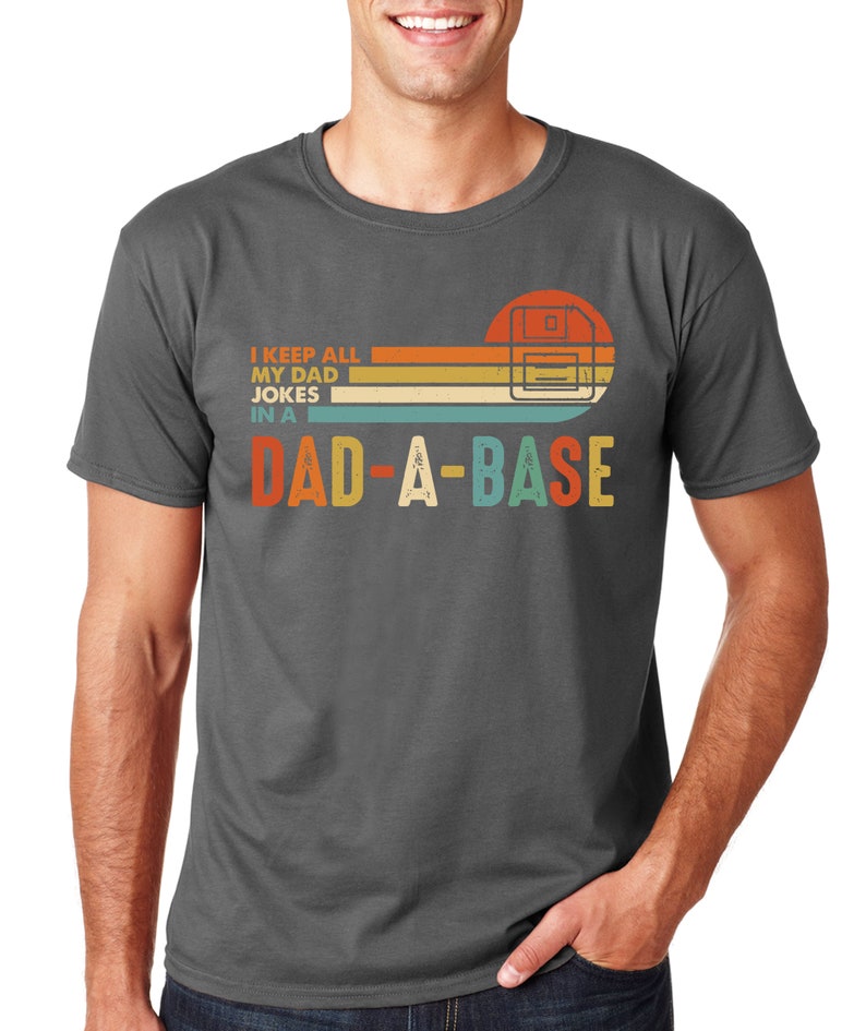 Op de afbeelding: Donkergrijs t-shirt met een retro design. Het ontwerp toont een floppy disk graphic met de tekst "I KEEP ALL MY DAD JOKES IN A DAD-A-BASE" in oranje, geel, blauwgroen en wit. Het t-shirt heeft een ronde hals en korte mouwen.