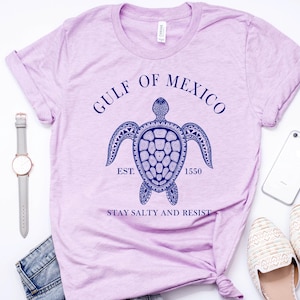 Puede incluir: Camiseta de color lila claro con un dise&ntilde;o de tortuga azul marino y el texto "GULF OF MEXICO EST. 1550 STAY SALTY AND RESIST". La camiseta se presenta con un reloj, vaqueros, un tel&eacute;fono y zapatos.