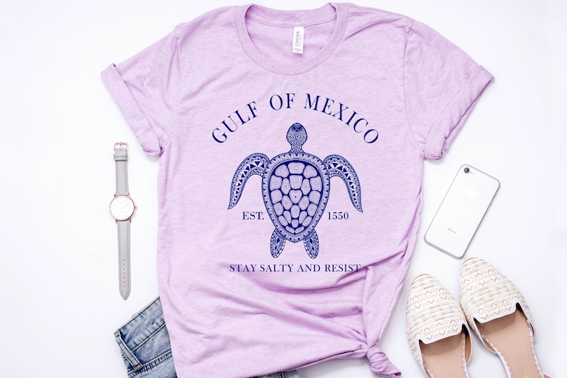 Puede incluir: Camiseta de color lila claro con un dise&ntilde;o de tortuga azul marino y el texto "GULF OF MEXICO EST. 1550 STAY SALTY AND RESIST". La camiseta se presenta con un reloj, vaqueros, un tel&eacute;fono y zapatos.