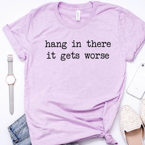 May include: A light purple t-shirt with the text "hang in there it gets worse" in black. The shirt is styled with a gray watch, denim shorts, a white phone, and cream-colored flats. The items are arranged on a white surface.