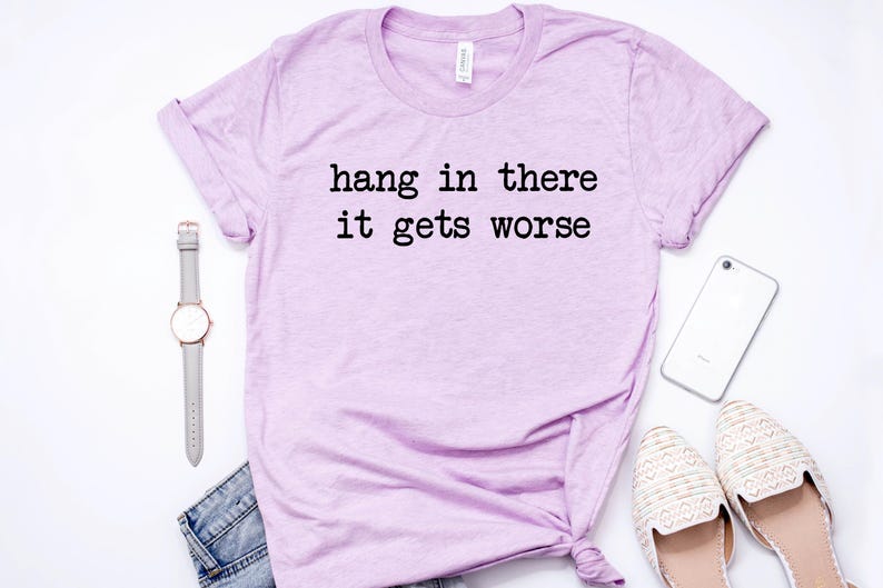 May include: A light purple t-shirt with the text "hang in there it gets worse" in black. The shirt is styled with a gray watch, denim shorts, a white phone, and cream-colored flats. The items are arranged on a white surface.