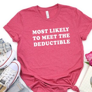 May include: A red t-shirt with the text "MOST LIKELY TO MEET THE DEDUCTIBLE" in white. The shirt is paired with blue jeans, white sneakers, and a gift bag with pink roses. A casual, everyday outfit.