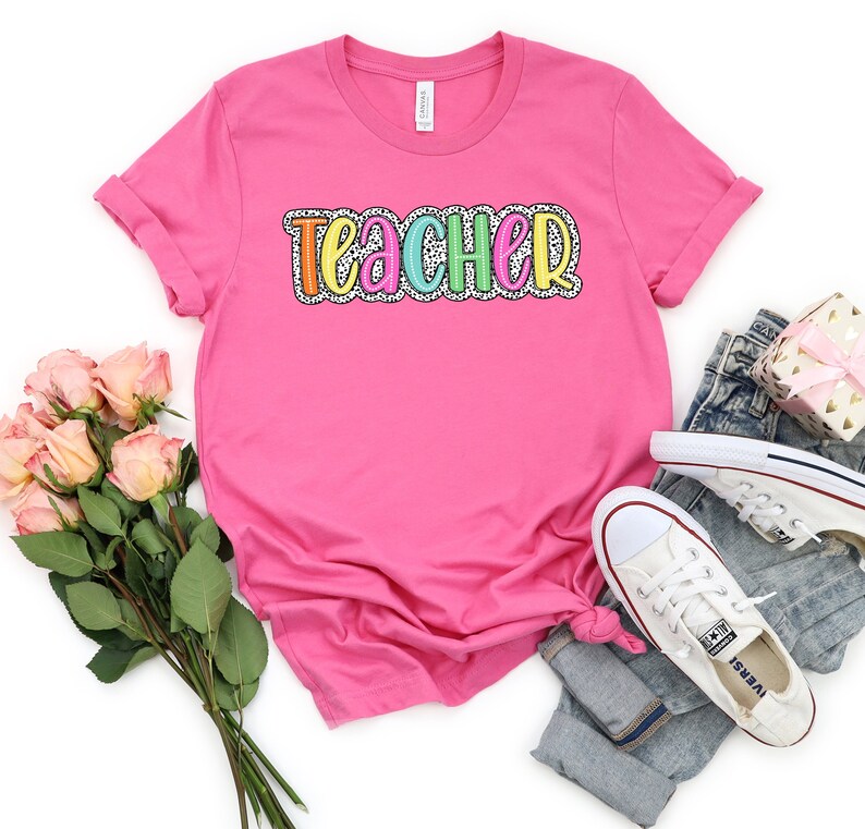 Puede incluir: Camiseta rosa con la palabra "TEACHER" en colores arco&iacute;ris con un patr&oacute;n de lunares blancos.