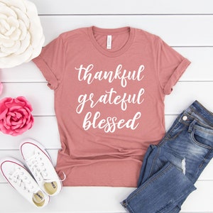 May include: A dusty rose colored t-shirt with the words "thankful grateful blessed" written in white on the front. The shirt is laying on a white wooden surface with a pair of white sneakers, blue jeans, and pink paper flowers.