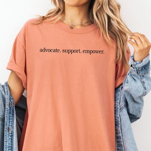 Op de afbeelding: Een perzikkleurig T-shirt met de woorden "advocate. support. empower." in het zwart gedrukt. Het shirt heeft korte mouwen en een ronde hals. Een spijkerjasje is over de schouders gedrapeerd.