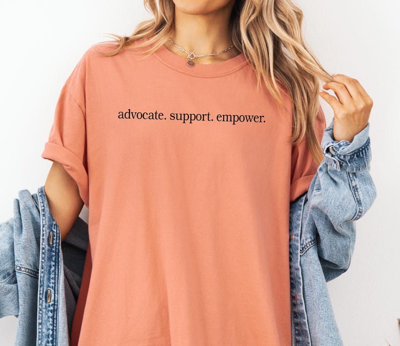 Op de afbeelding: Een perzikkleurig T-shirt met de woorden "advocate. support. empower." in het zwart gedrukt. Het shirt heeft korte mouwen en een ronde hals. Een spijkerjasje is over de schouders gedrapeerd.