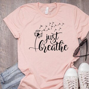 Op de afbeelding: Een roze T-shirt met een zwarte paardenbloem afbeelding en de tekst "just breathe".