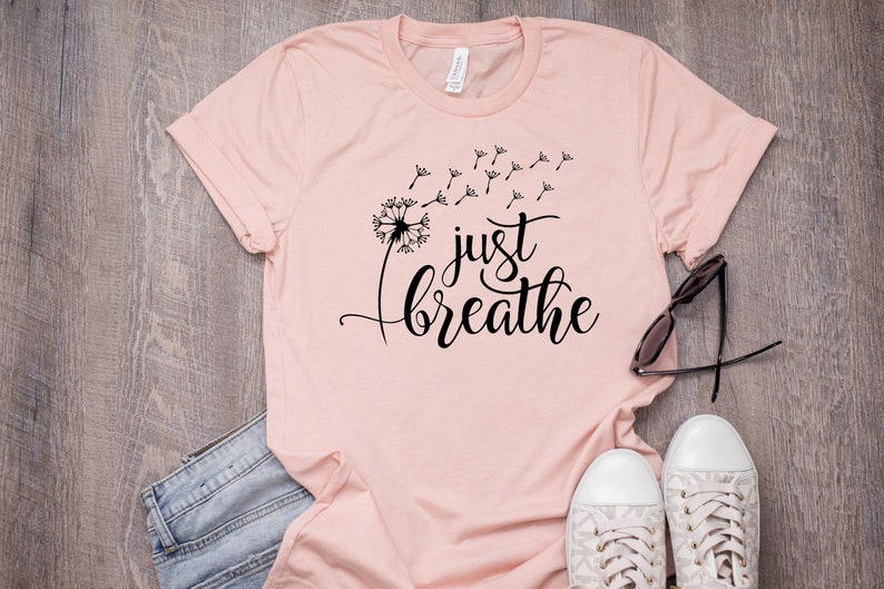 Op de afbeelding: Een roze T-shirt met een zwarte paardenbloem afbeelding en de tekst "just breathe".