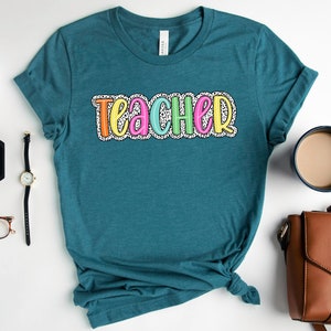 Puede incluir: Camiseta azul turquesa con la palabra "teacher" en letras de colores con lunares blancos.