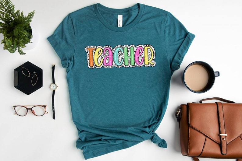 Puede incluir: Camiseta azul turquesa con la palabra "teacher" en letras de colores con lunares blancos.