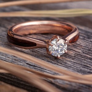 May include: A rose gold engagement ring with a round diamond set in a six-prong setting. The band is made of wood with a dark brown finish.