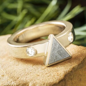 Puede incluir: Un anillo de color dorado con una incrustación triangular de piedra gris. El anillo presenta dos pequeñas piedras preciosas transparentes en la banda. La piedra triangular tiene un patrón texturizado único. El anillo está sobre una superficie marrón claro.