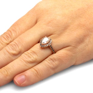 May include: A pear-shaped diamond engagement ring set in rose gold with a halo of black diamonds.