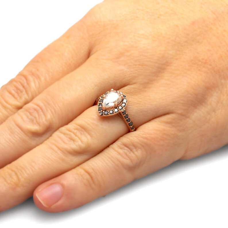 May include: A pear-shaped diamond engagement ring set in rose gold with a halo of black diamonds.