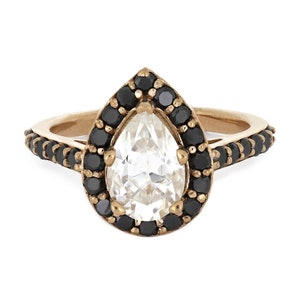 May include: A pear-shaped diamond ring with a black diamond halo setting. The ring is made of gold and has a delicate band.