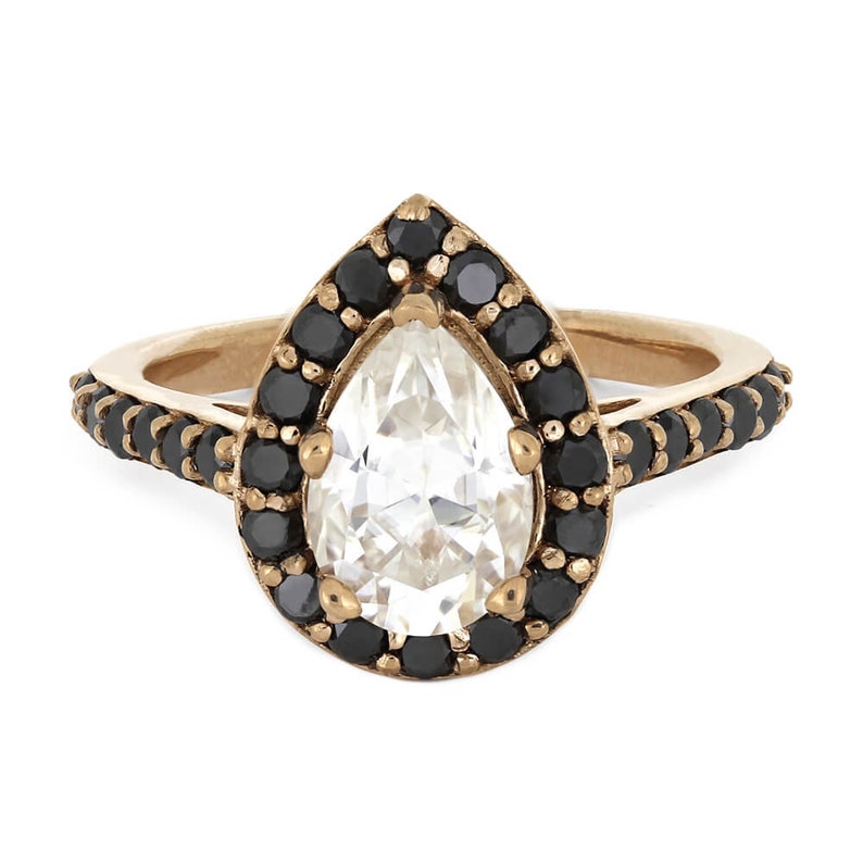 May include: A pear-shaped diamond ring with a black diamond halo setting. The ring is made of gold and has a delicate band.