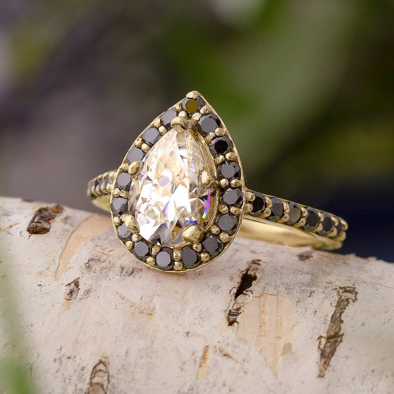 May include: A pear-shaped diamond engagement ring with a black diamond halo setting. The ring is set in yellow gold and features a delicate band.
