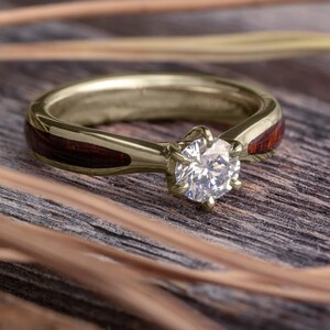 May include: A gold ring with a round, clear diamond set in a prong setting. The band of the ring is made of wood with a dark brown finish.