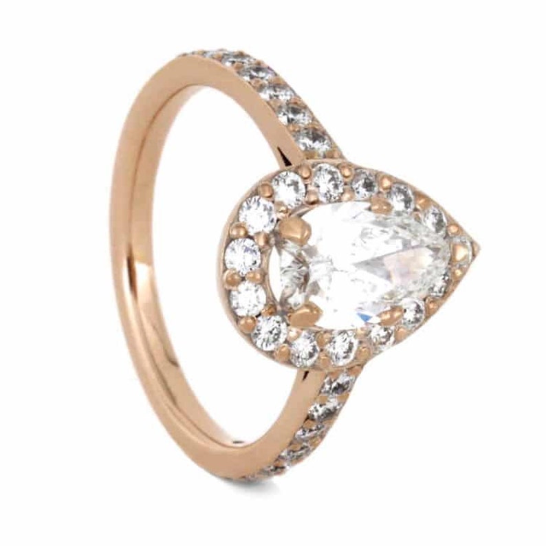 May include: A pear-shaped diamond engagement ring set in rose gold with a halo of smaller diamonds. The band is also set with smaller diamonds.