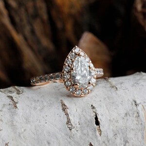 May include: A pear-shaped diamond engagement ring with a halo setting in rose gold. The ring has a band of small diamonds.