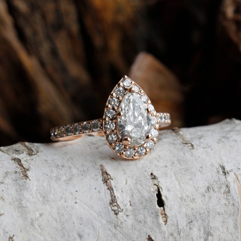 May include: A pear-shaped diamond engagement ring with a halo setting in rose gold. The ring has a band of small diamonds.