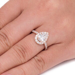 May include: A pear-shaped diamond engagement ring set in rose gold with a halo of smaller diamonds. The ring is on a finger.