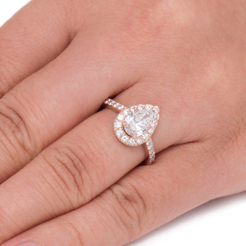 May include: A pear-shaped diamond engagement ring set in rose gold with a halo of smaller diamonds. The ring is on a finger.