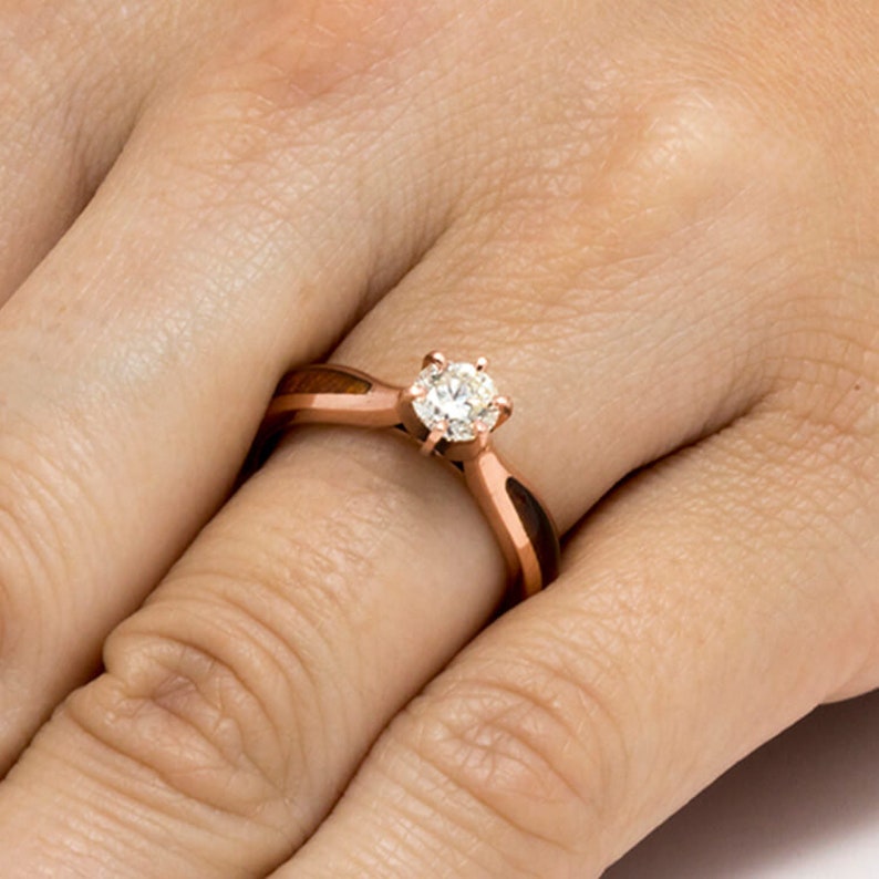 May include: A rose gold engagement ring with a round diamond set in a simple prong setting. The band is made of wood with a brown and black inlay.