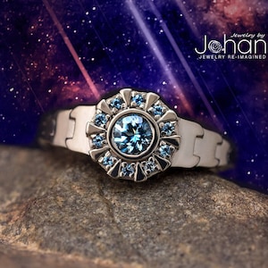 Może przedstawiać: Srebrny pierścionek z dużym niebieskim kamieniem szlachetnym otoczonym mniejszymi niebieskimi kamieniami. Pierścionek leży na szarym kamieniu na tle rozgwieżdżonego nieba.