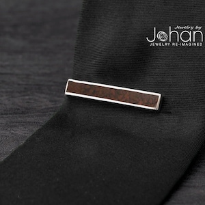 May include: A silver tie bar with a brown wood inlay. The tie bar is on a black tie. The text "Jewelry by Johan Jewelry Re-Imagined" is in the top right corner of the image.