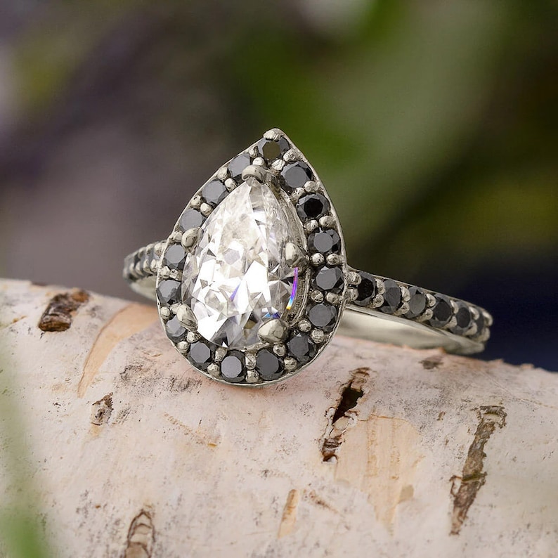 May include: A pear-shaped diamond engagement ring with a black diamond halo setting. The ring is set in silver and has a delicate band.