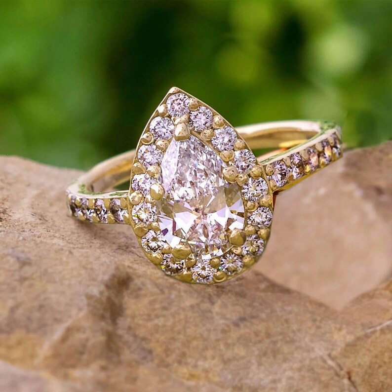 May include: A pear-shaped diamond engagement ring with a halo setting. The ring is set in yellow gold and features a row of smaller diamonds on the band.