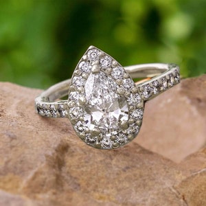 May include: A pear-shaped diamond engagement ring with a halo setting. The ring is set in white gold and features a row of smaller diamonds on the band.