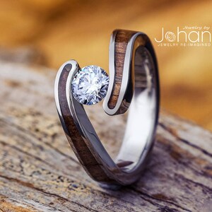 Puede incluir: Un anillo único con un diamante brillante de talla redonda engastado en un diseño en espiral. El anillo combina madera y metal plateado, con el texto "Jewelry by Johan" visible en el fondo.