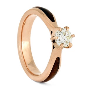 May include: A rose gold engagement ring with a round diamond set in a four-prong setting. The band is made of wood with a black inlay.