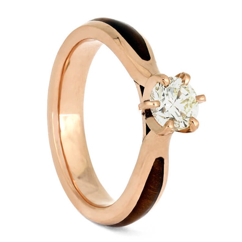 May include: A rose gold engagement ring with a round diamond set in a four-prong setting. The band is made of wood with a black inlay.