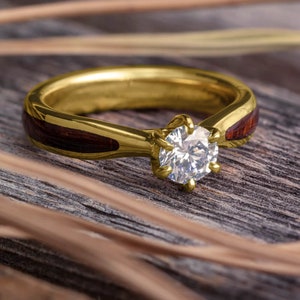 May include: A gold ring with a single round diamond set in a prong setting. The band of the ring is inlaid with a thin strip of dark wood.