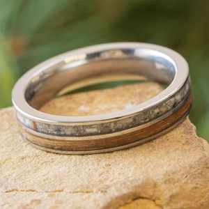 May include: A close-up of a silver ring with a unique design. The ring features a band of light-colored stone and a band of wood-like material, set between silver edges. The ring is displayed on a light brown surface.