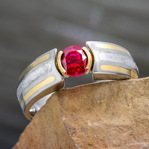 May include: A silver and gold ring with a large, round, red gemstone. The ring has a unique design with gold accents and a textured silver band. The gemstone is set in a gold bezel, creating a striking contrast.