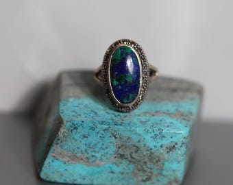 May include: A silver ring with a blue and green oval gemstone set in a silver setting with a decorative border. The ring is sitting on a turquoise surface.